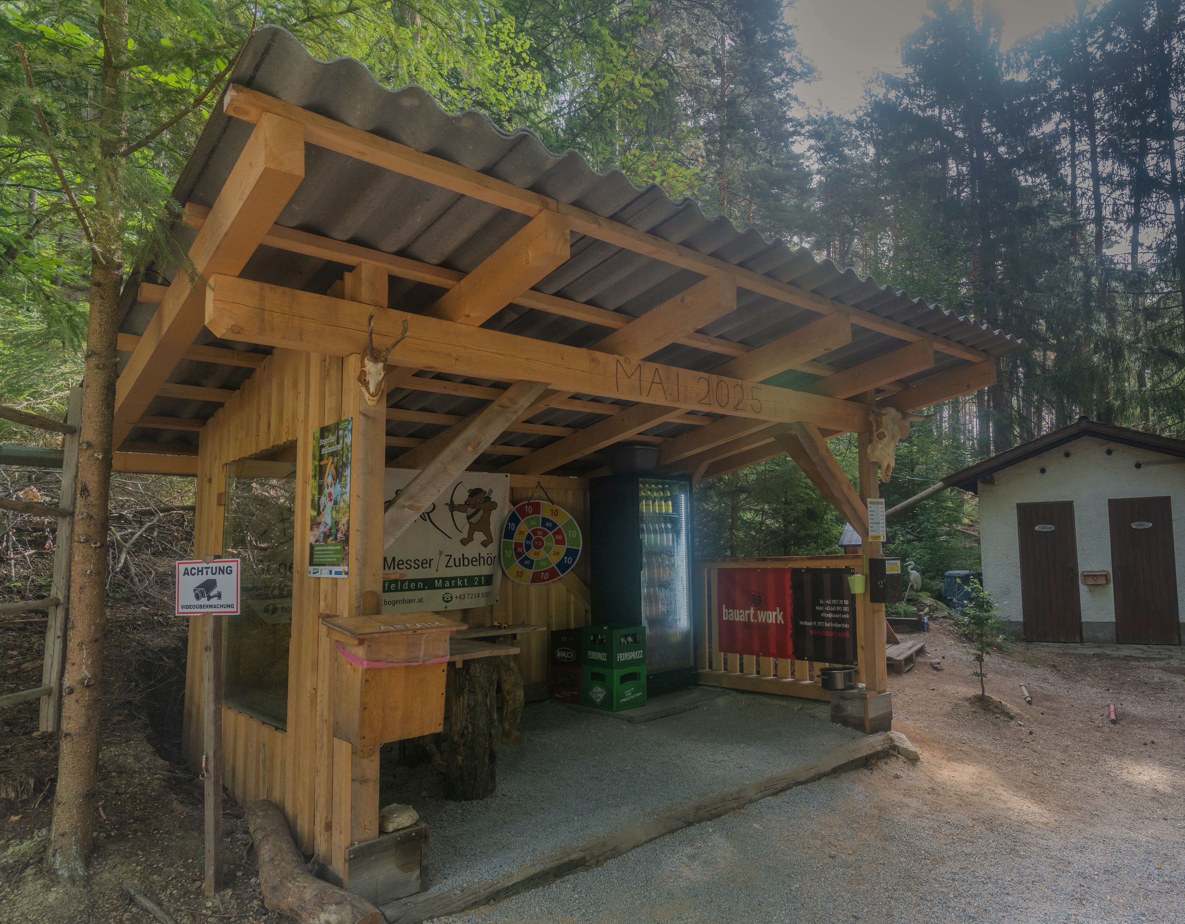 Überdachte Labstelle aus Holz mit Sitzgelegenheiten, Getränkekühlschrank und Zielscheibe am Bogenparcours im Naturpark Nordwald